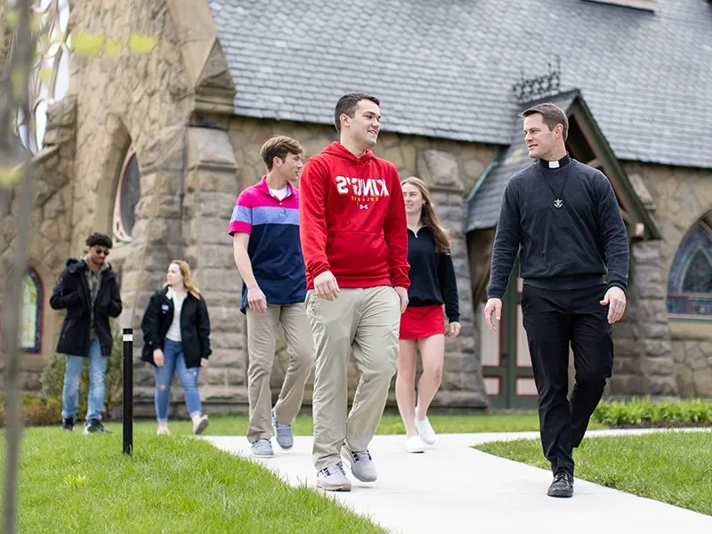 a priest walks with several students outside the Chapel
